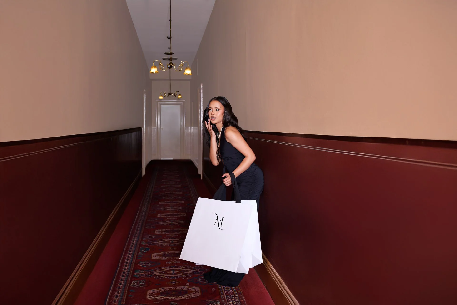 A woman in a black dress holding shopping bags in a hallway with beige walls and red carpet.
