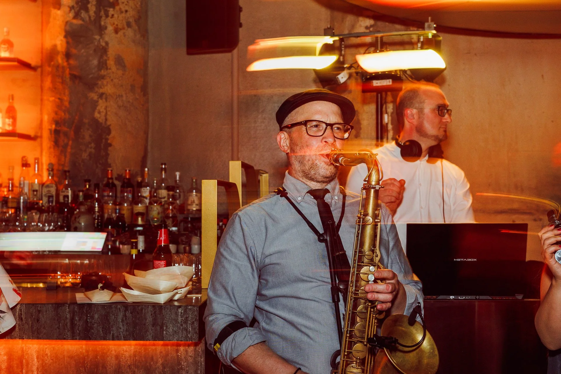 A man playing a saxophone in a bar with bottles of alcohol on the shelves behind him, another man wearing headphones standing nearby, dim warm lighting, exposed brick wall, and a bar counter with napkins and condiments.
