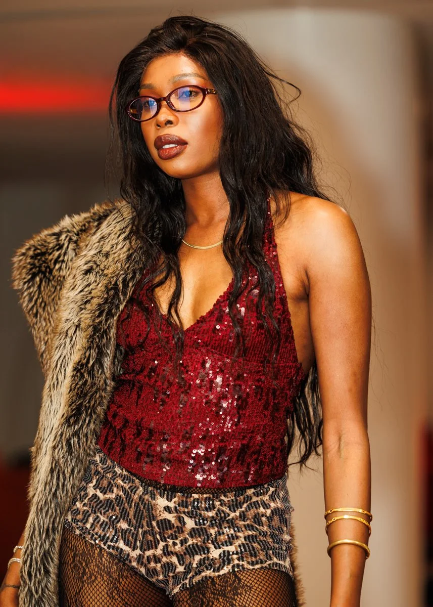 A woman with dark wavy hair wearing glasses, a red sequined top, leopard print shorts, fishnet stockings, and a faux fur coat draped over one shoulder.