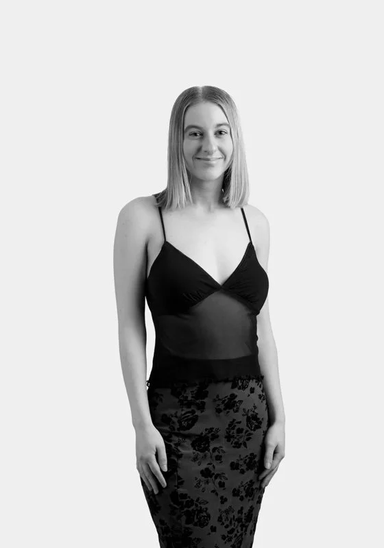 Black and white photo of a young woman with shoulder-length hair, smiling, wearing a spaghetti strap top and a floral skirt.