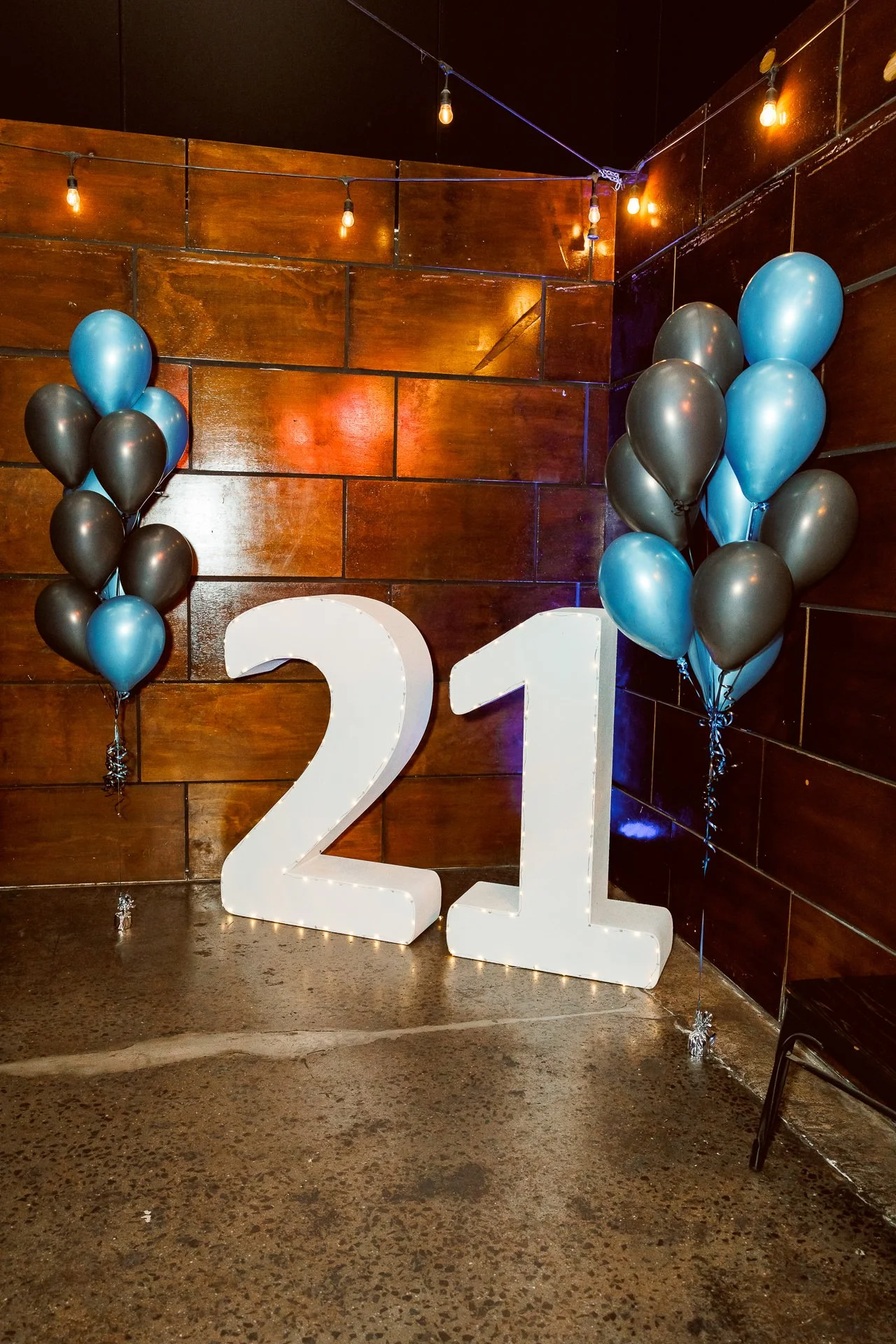 Decorative 21st birthday celebration with large white number "21" sign, black and blue balloons on either side, and string lights hanging overhead against a dark wood-paneled wall.