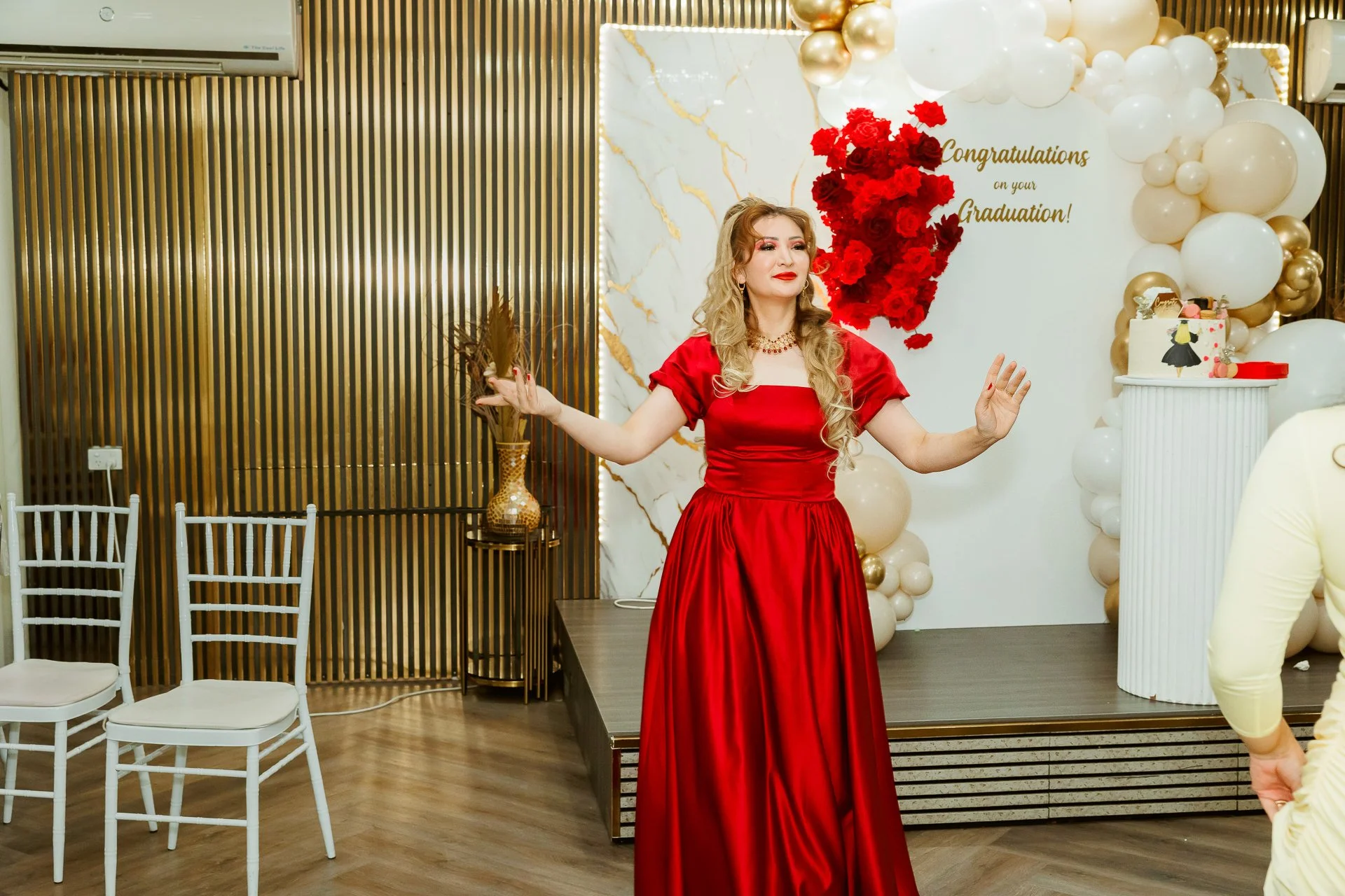 A woman in a red dress standing with arms outstretched at a graduation celebration, with a decorated backdrop featuring balloons, red flowers, a cake, and a congratulatory message.