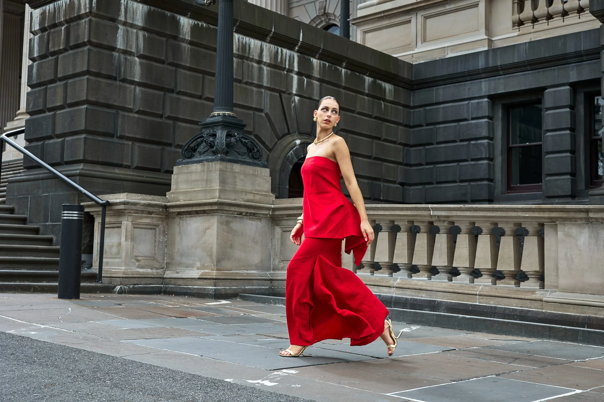 A woman in a red strapless dress and high heels walking on city sidewalk with historic building in background.