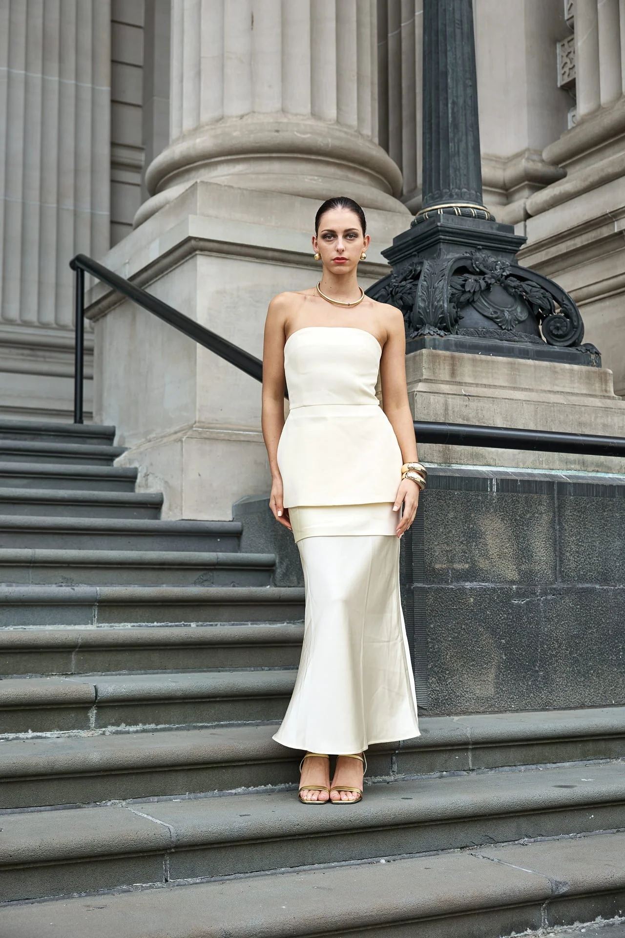 A woman in an elegant, strapless white dress standing on stone stairs outside a historic building with tall columns and ornate details, accessorized with gold jewelry.