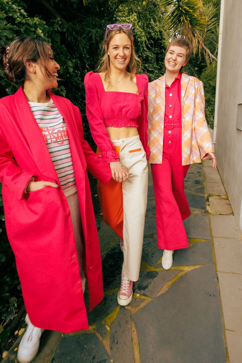 Three women walking outdoors, holding hands and smiling, dressed in bold, colorful outfits with pink and orange hues.