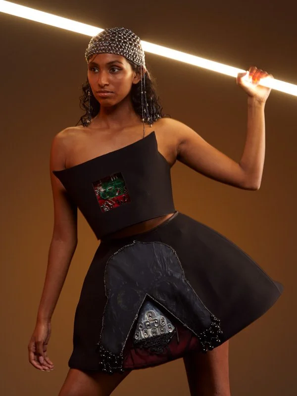 A woman wearing a black asymmetrical dress with electronic circuit details, a silver beaded headpiece, and holding a horizontal glowing light tube.