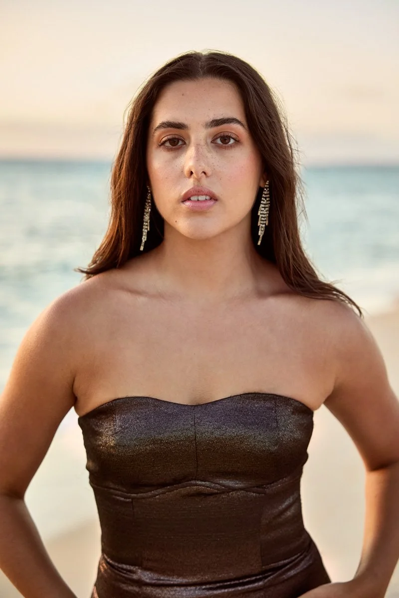A young woman with dark brown hair, fair skin, and wearing a strapless dark dress and long earrings, standing outdoors near the water at sunset.