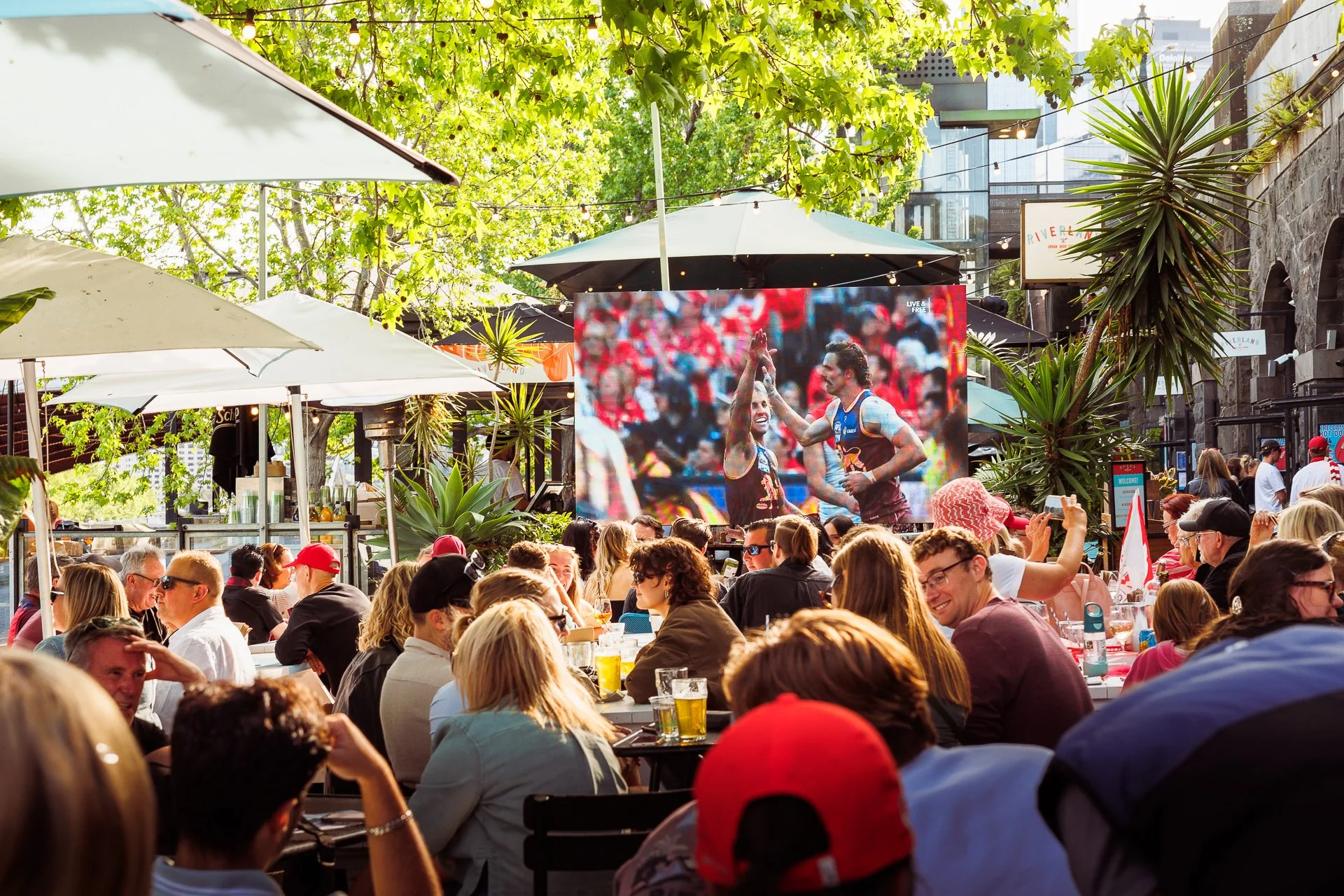 Crowd of people watching outdoor screen showing a sports event, surrounded by trees, umbrellas, and plants in a lively patio or outdoor bar area.