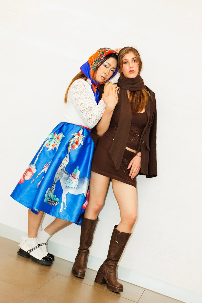 Two women standing against a white wall, one dressed in a colorful costume with a patterned headscarf and skirt, the other wearing a brown outfit with knee-high boots.