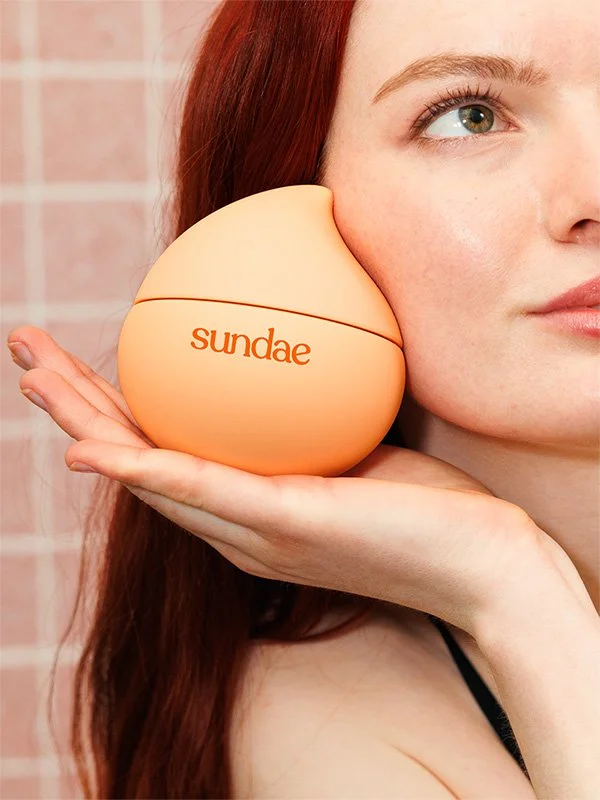 A woman with red hair holding a round, orange container with the word "sundae" written on it, resting on her cheek.