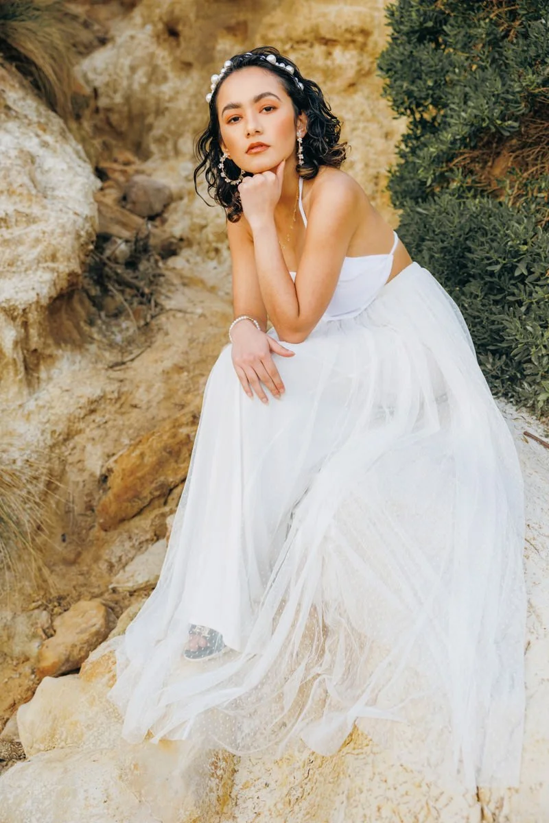 A young woman in a white dress and jewelry sitting on rocks outdoors with desert-like surroundings and greenery.