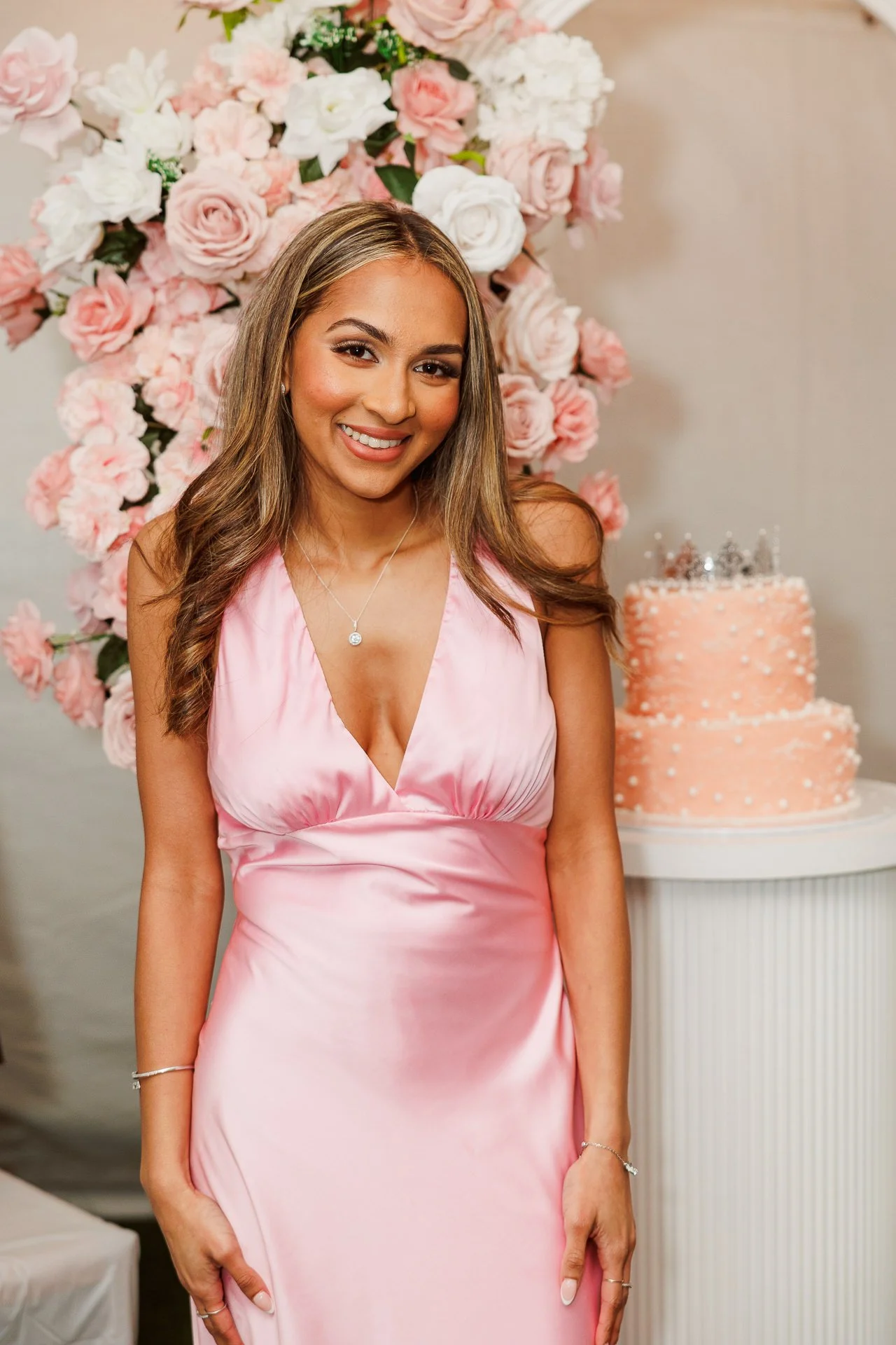 A young woman with long wavy hair wearing a pink satin dress with a deep neckline, smiling at the camera. Behind her is a large floral display of pink and white roses, and to her right is a peach-colored, two-tiered cake decorated with white dots and
