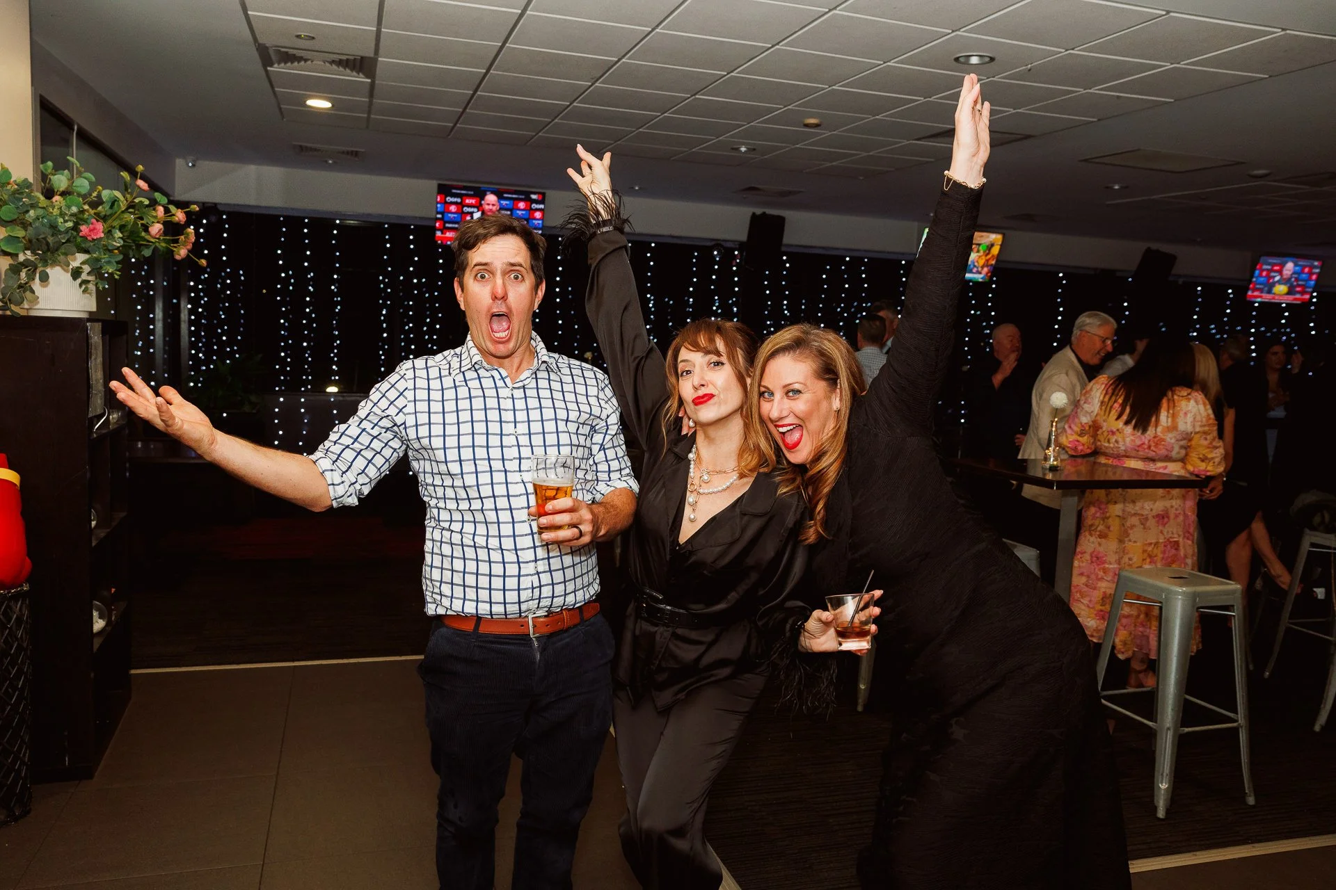 Three friends having a good time at a party. The man on the left holds a glass of beer, the woman in the middle has her arm raised and is dressed in black, and the woman on the right is smiling, holding a glass with a drink. They are all smiling and 