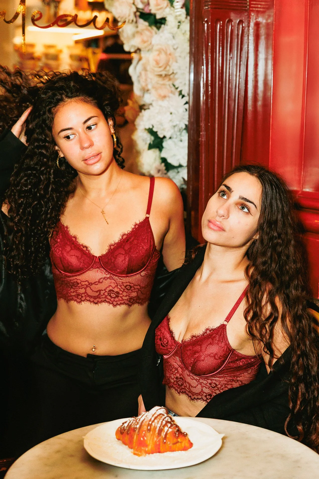 Two women with curly hair, wearing matching red lace bralettes and black jackets, posing in a decorated indoor setting with flowers and a dessert on a table in front of them.