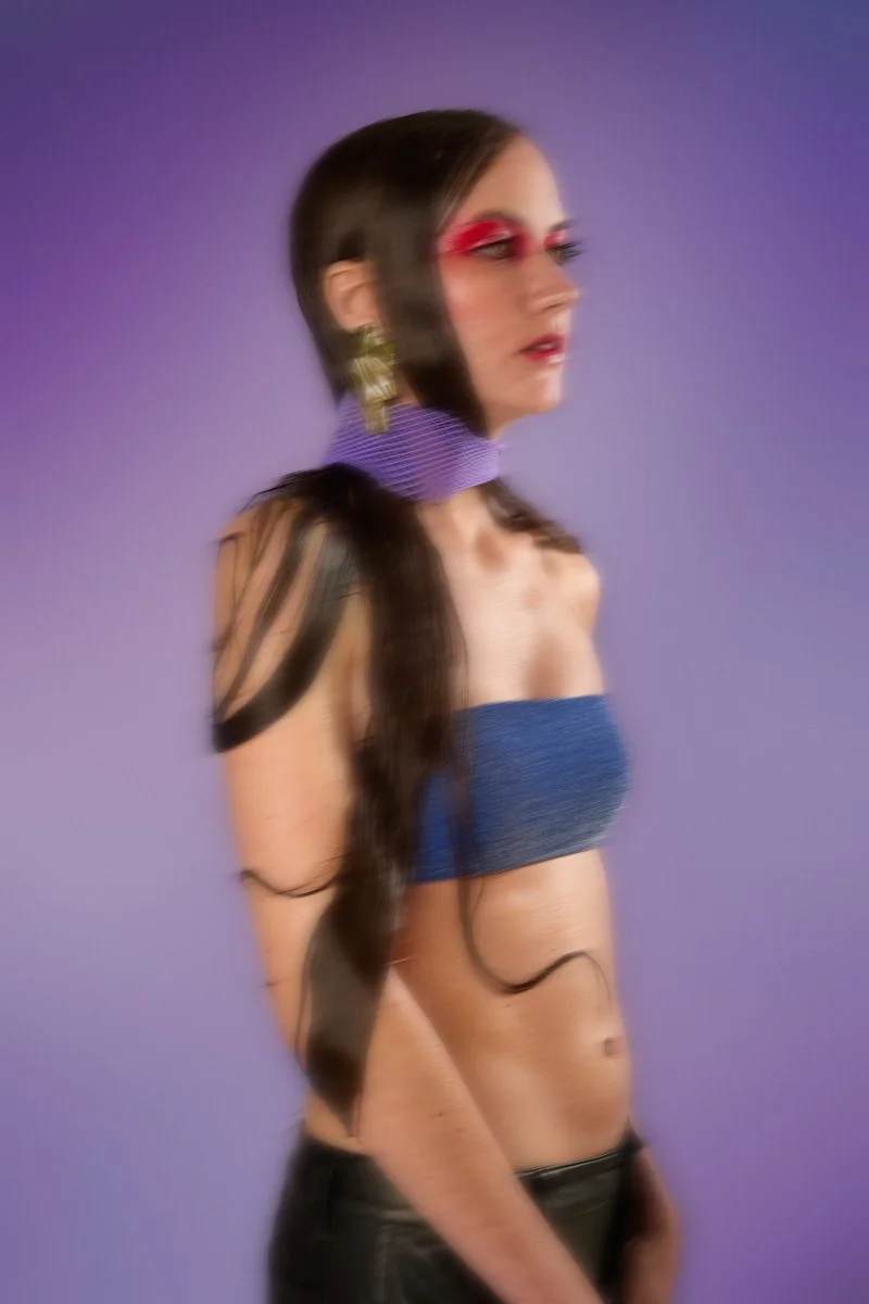 A woman with long dark hair, wearing bold red glasses, a purple choker, a strapless blue top, earrings, and black pants, standing against a purple background.
