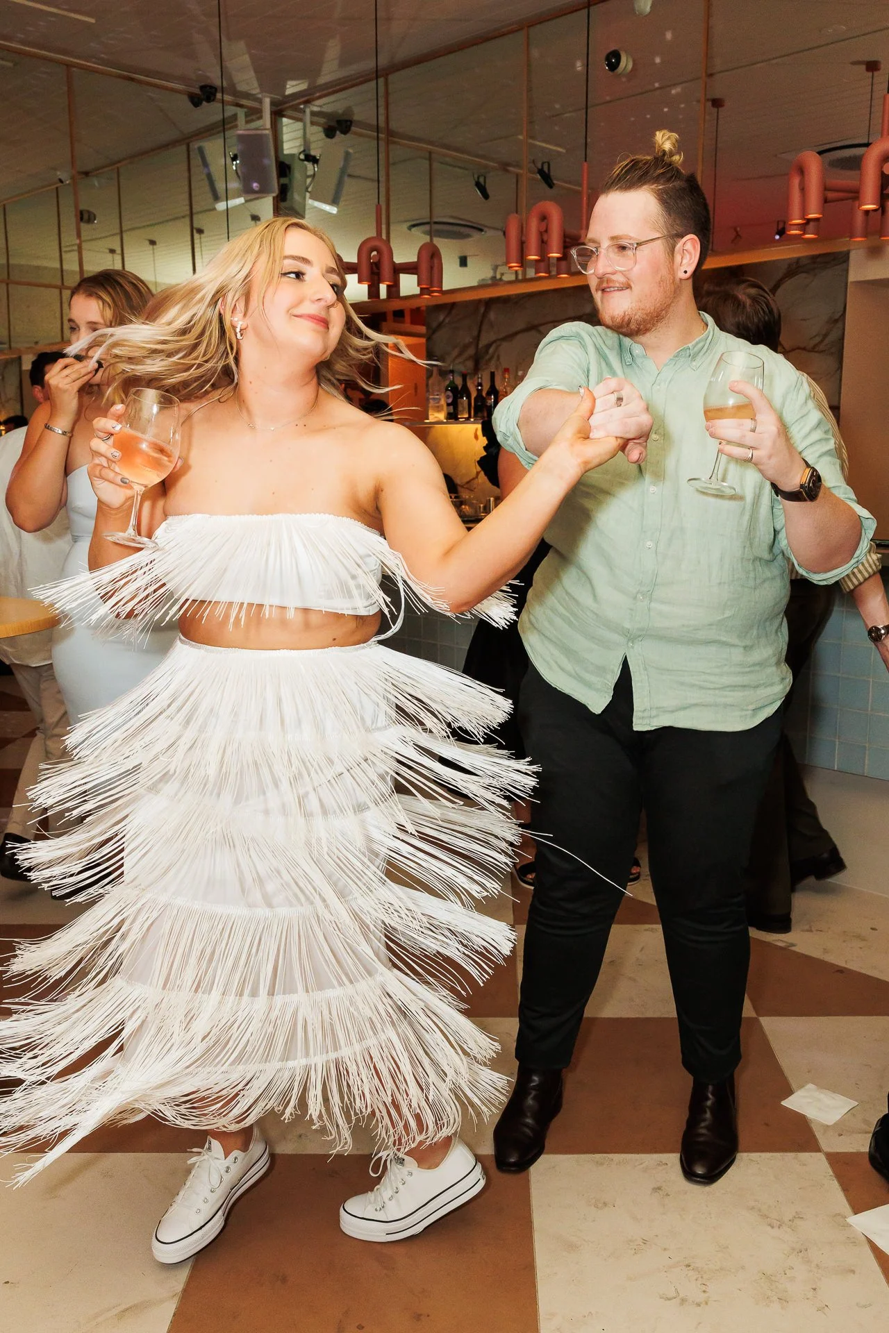 A woman in a white, fringed dress dancing and holding a glass of wine, while a man in a light green shirt and black pants dances with her, holding her hand and also holding a glass of wine. They are in a lively indoor setting with other people in the