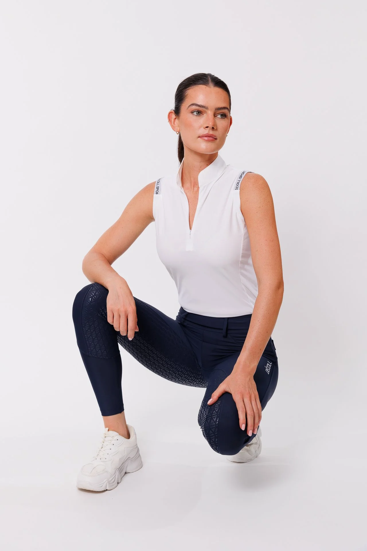 A woman in athletic clothing crouching, wearing a white sleeveless top and navy-blue leggings with a geometric pattern, against a plain white background.