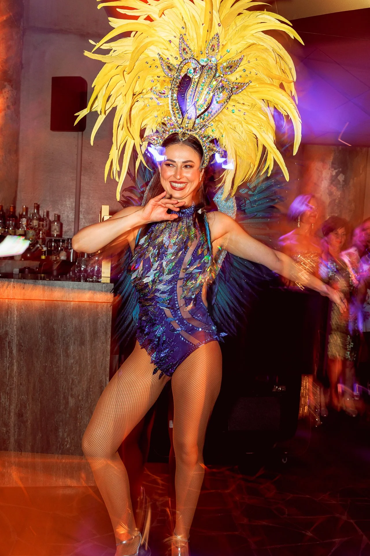 A woman dressed in a colorful show girl costume with a large yellow feather headdress, posing and smiling at a lively indoor event.