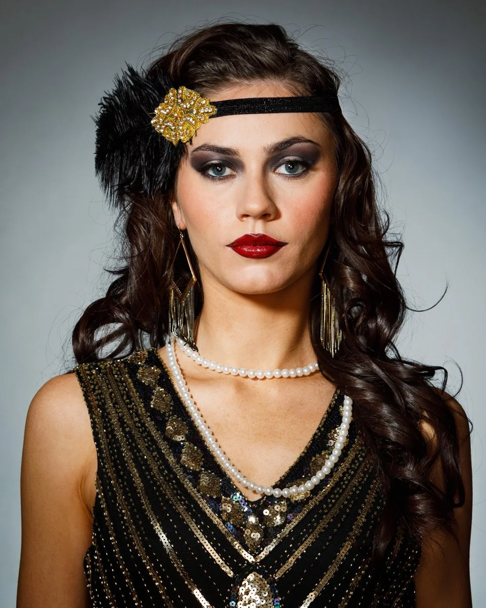 A woman with dark wavy hair wearing vintage style black dress with gold and sequin embellishments, a pearl necklace, gold dangling earrings, a black headband with a gold flower and feather decoration, and bold makeup including dark eyeshadow and red 