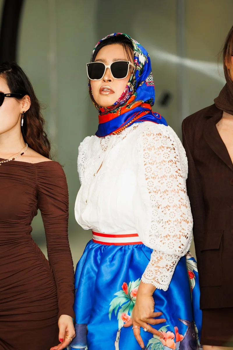 A woman wearing a colorful headscarf, large sunglasses, a white lace blouse, and a floral blue skirt, standing between two women in brown and black outfits.
