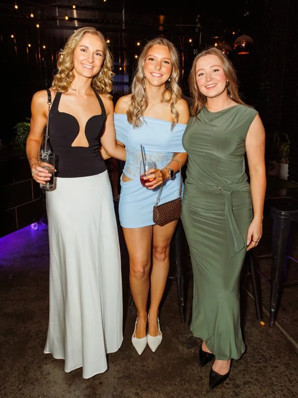 Three young women posing together at a party, all smiling and holding drinks, dressed in elegant evening attire.