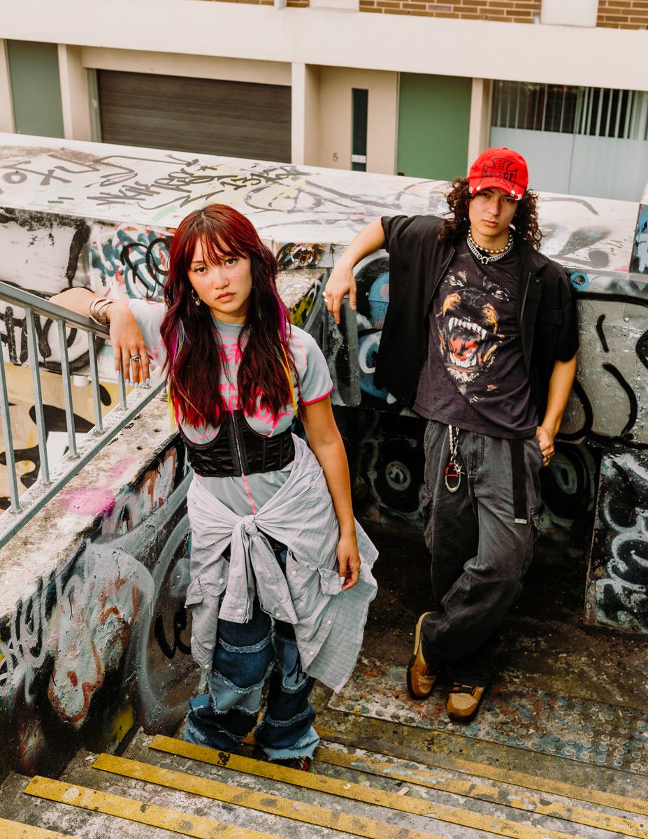 Two young individuals with edgy fashion styles posing against a graffiti-covered concrete staircase and wall in an urban setting. The girl on the left has long, wavy hair with pink highlights, wearing a graphic t-shirt, layered clothing, and distress