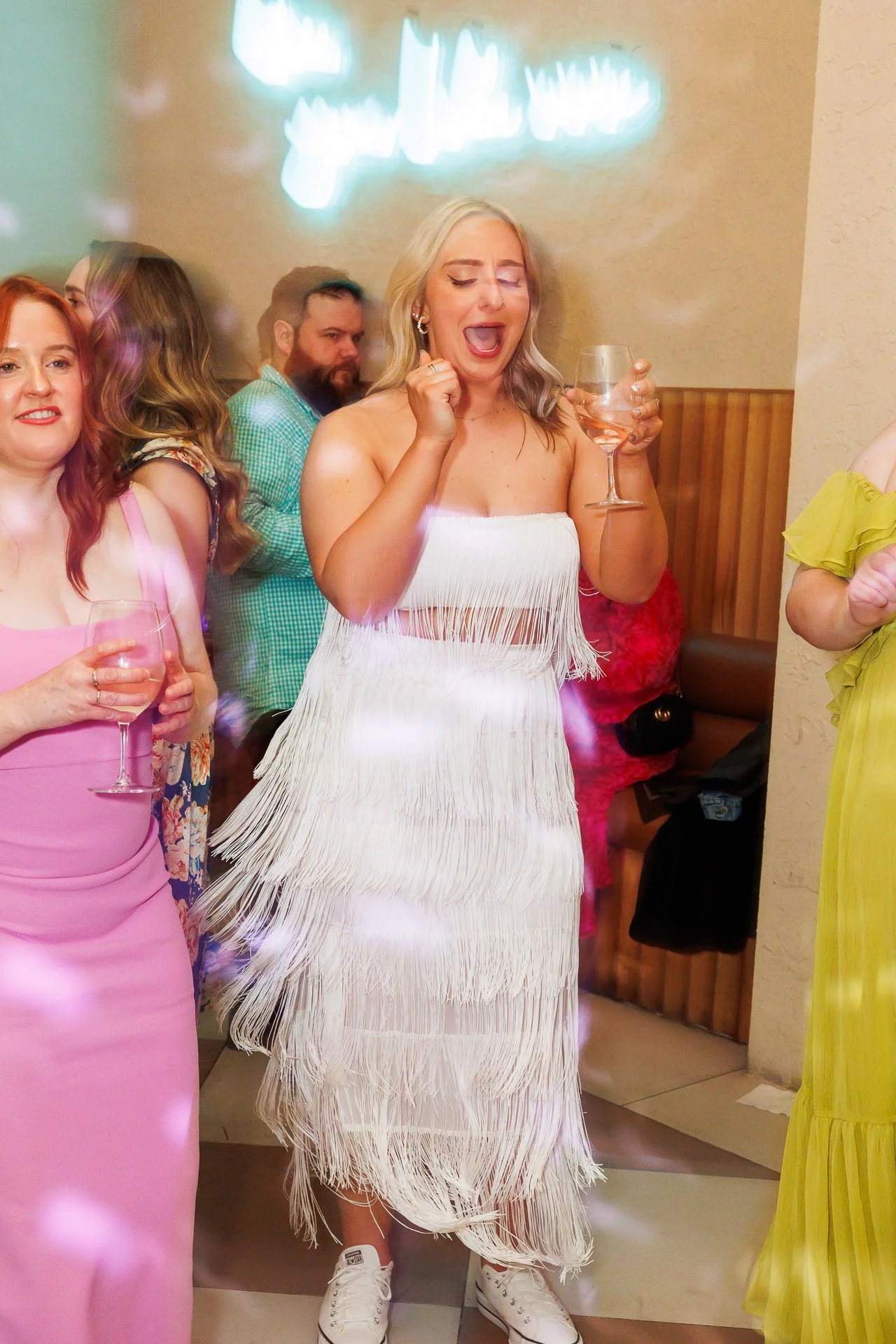 A woman in a white fringed dress holding a wine glass, singing or shouting at a celebration or party with other women and men, neon sign in background, indoors.