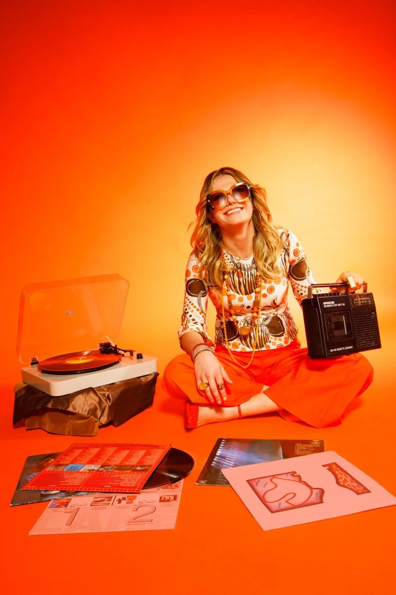 Woman with blonde hair wearing large sunglasses, colorful patterned top, and orange pants, sitting cross-legged on a bright orange background. She is holding a boombox in one hand and smiling. There is a turntable on her left, along with records and 