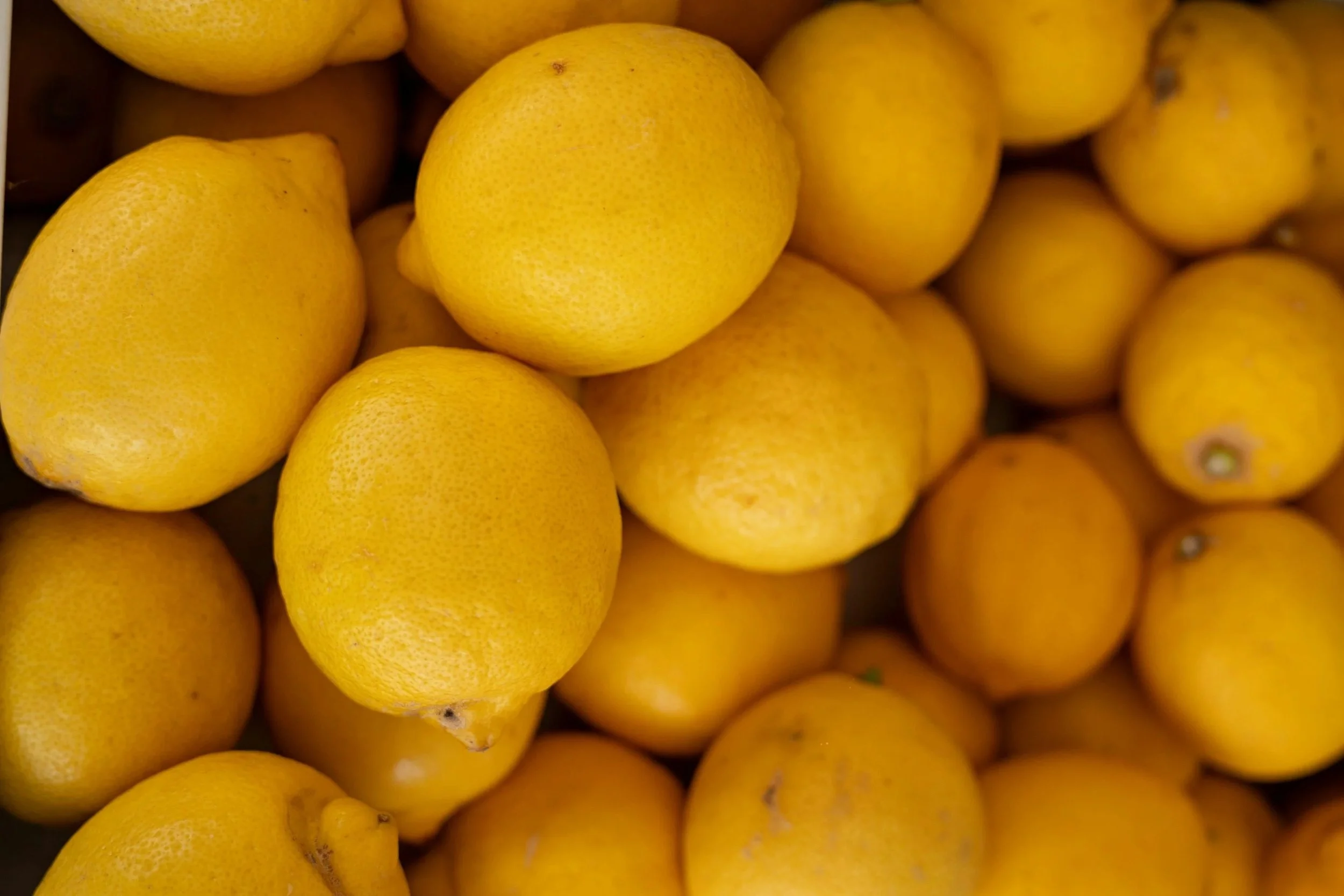 A close-up of numerous yellow lemons, some with slightly darker spots, piled together.