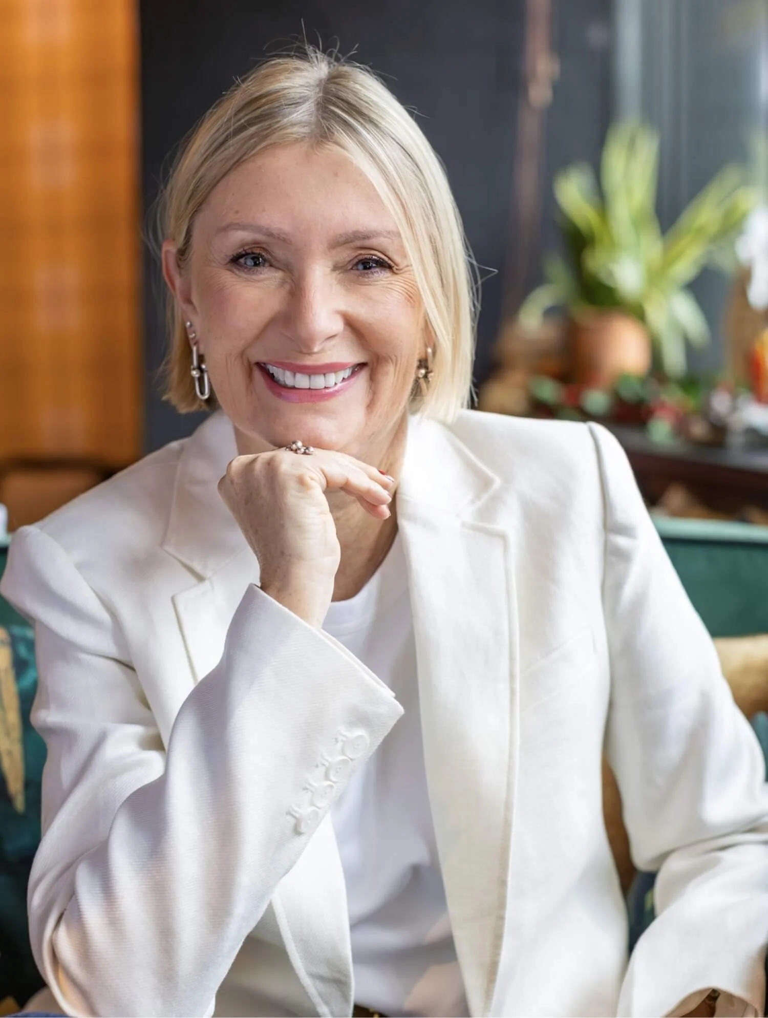 Smiling woman with blonde hair in a white blazer sitting in a cozy indoor space with plants and warm decor.