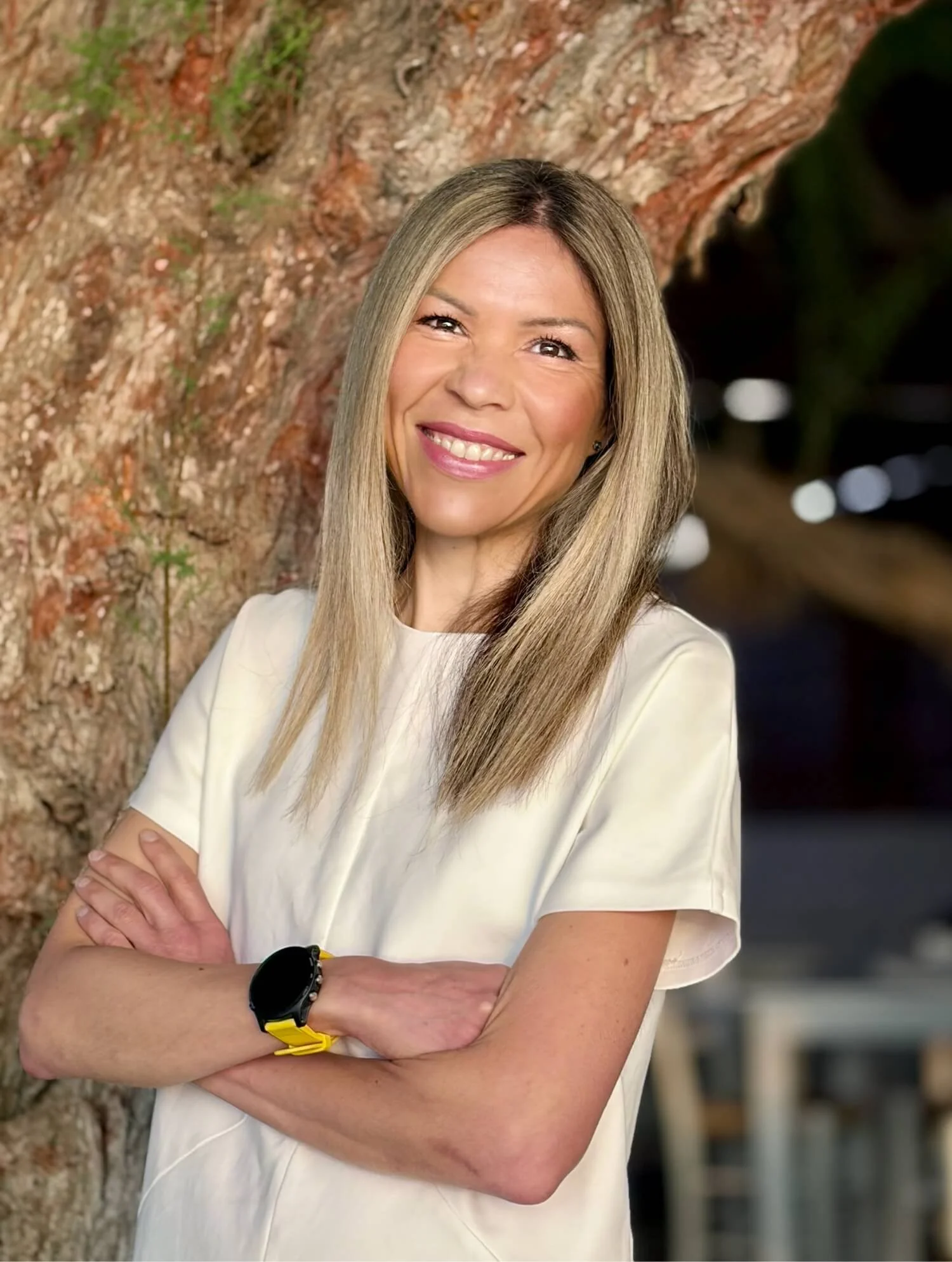 A woman with long blonde hair smiling and standing with arms crossed in front of a tree with rough bark.