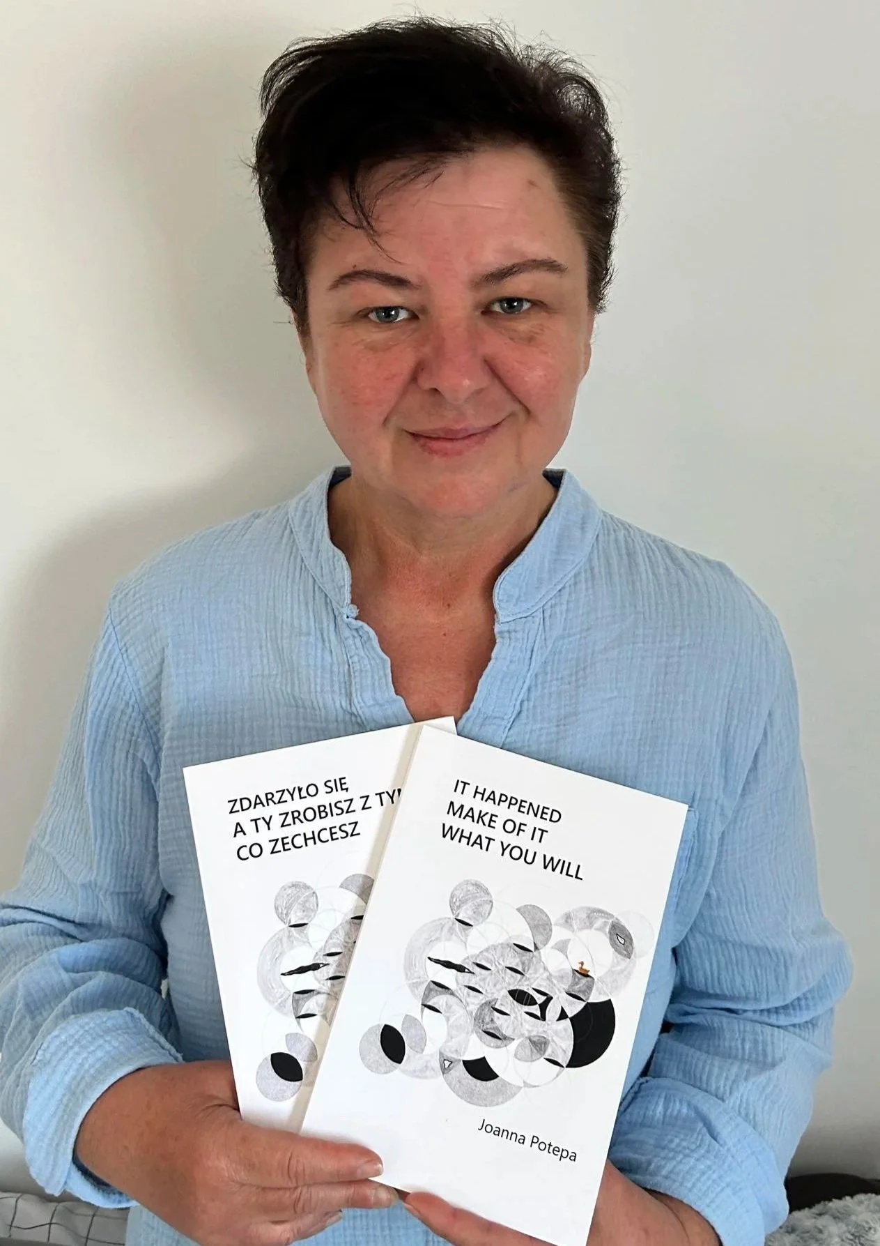 A woman with short dark hair and light skin holding two books titled 'It Happened Make of It What You Will' by Joanna Potepa, standing against a plain white wall, wearing a light blue button-up shirt.