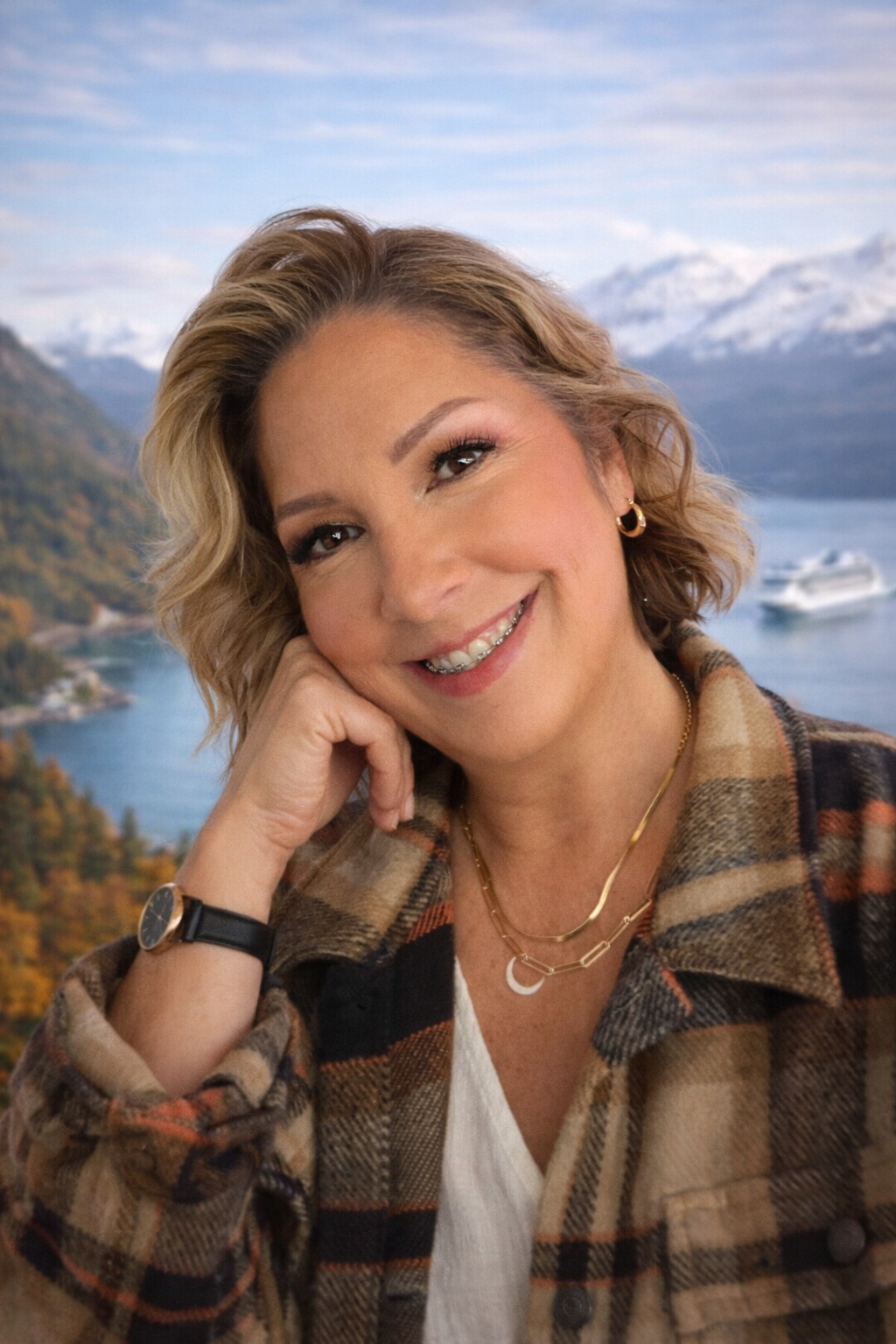 A woman with short wavy blonde hair smiling, wearing a plaid jacket, layered necklaces, and a watch, with a scenic background of mountains, water, and a boat.