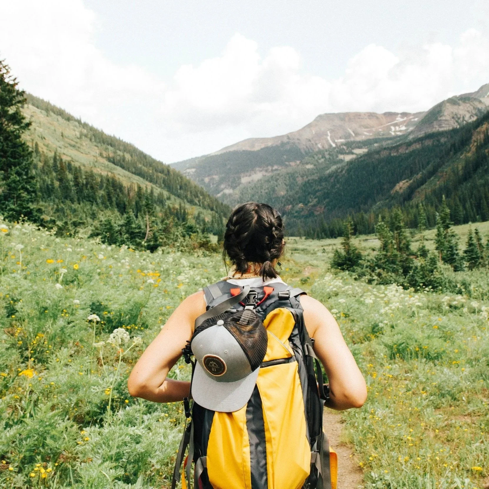 A woman hiking on a trail through a green valley surrounded by mountains, with her back to the camera and a gray and yellow backpack and cap.