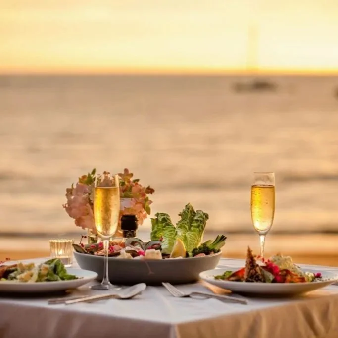 A dinner table set with plates of food, two glasses of champagne, and a centerpiece of flowers and vegetables by the ocean at sunset.