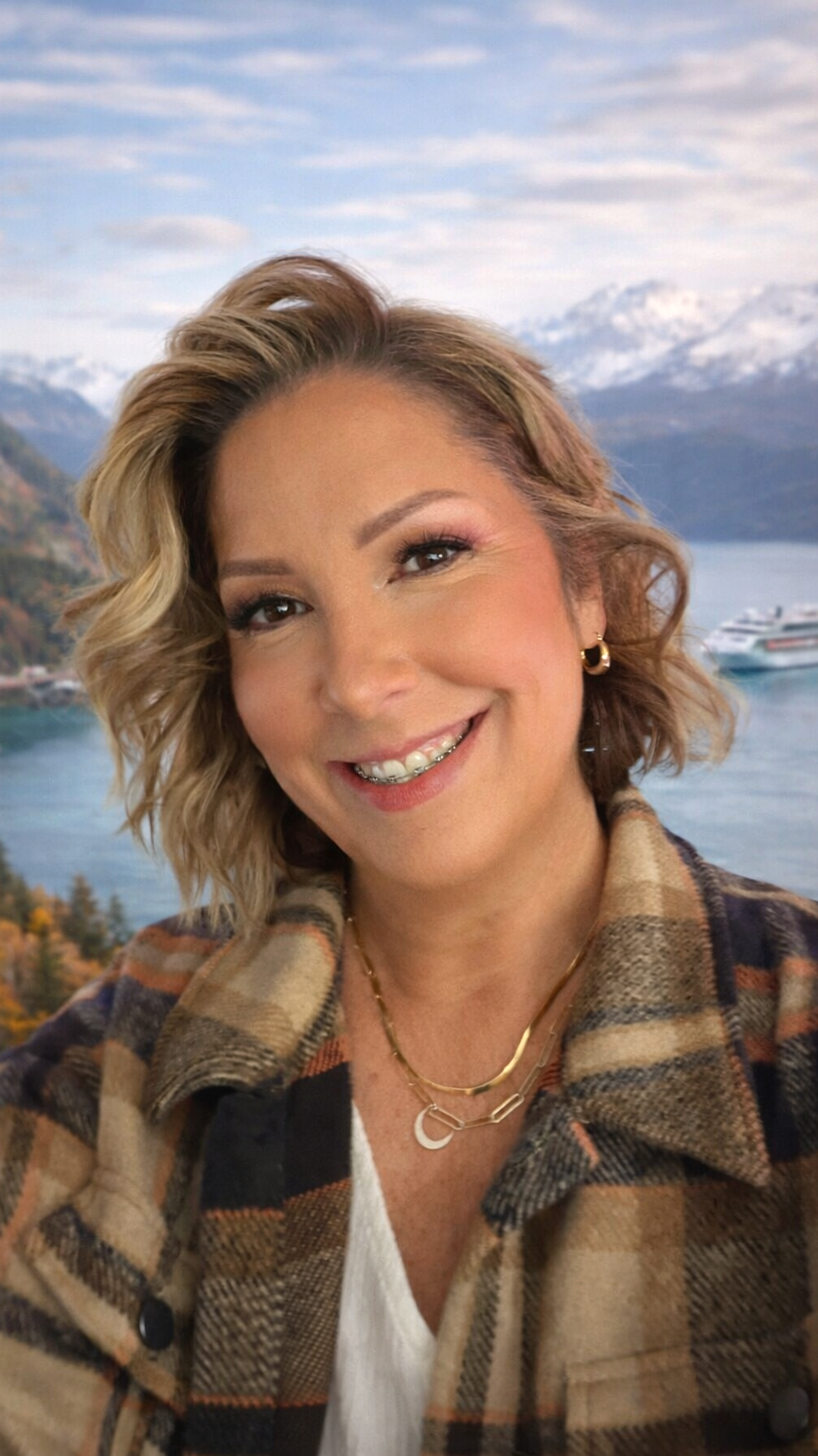 A woman with short, wavy blonde hair smiling, with a background of mountains, water, and a cruise ship.