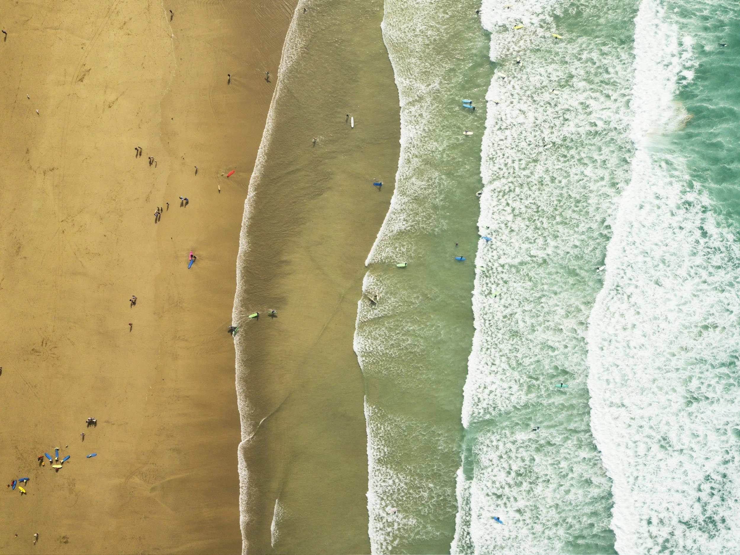 Aerial view of a sandy beach with people and colorful umbrellas, and ocean waves with surfers in the water.