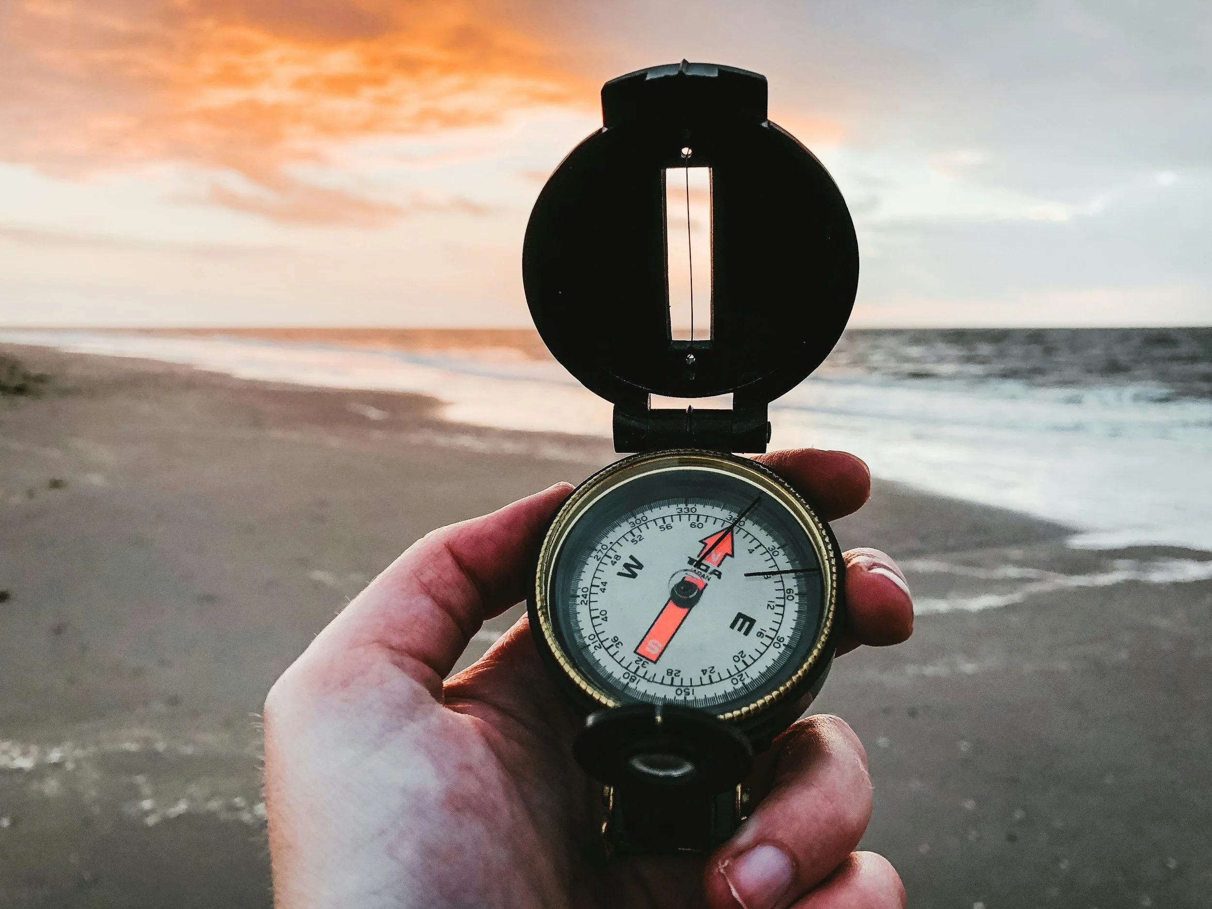 A person holding a compass on a beach during sunset or sunrise, with the ocean and sky in the background.