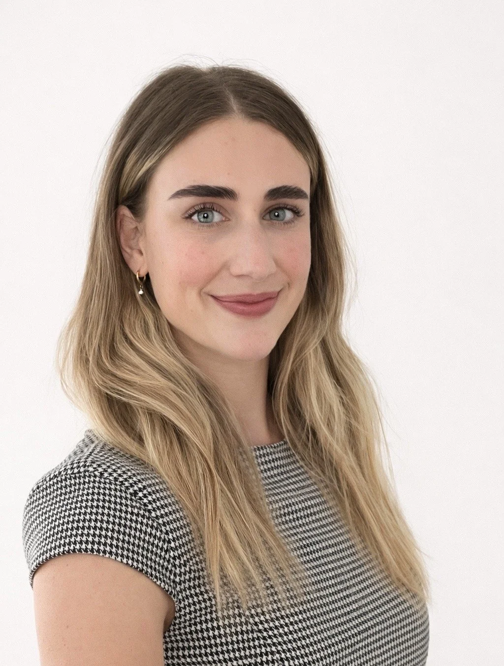 Portrait of a young woman with long, wavy blonde hair, blue eyes, and light makeup, wearing a black and white houndstooth pattern top, standing against a plain white background.