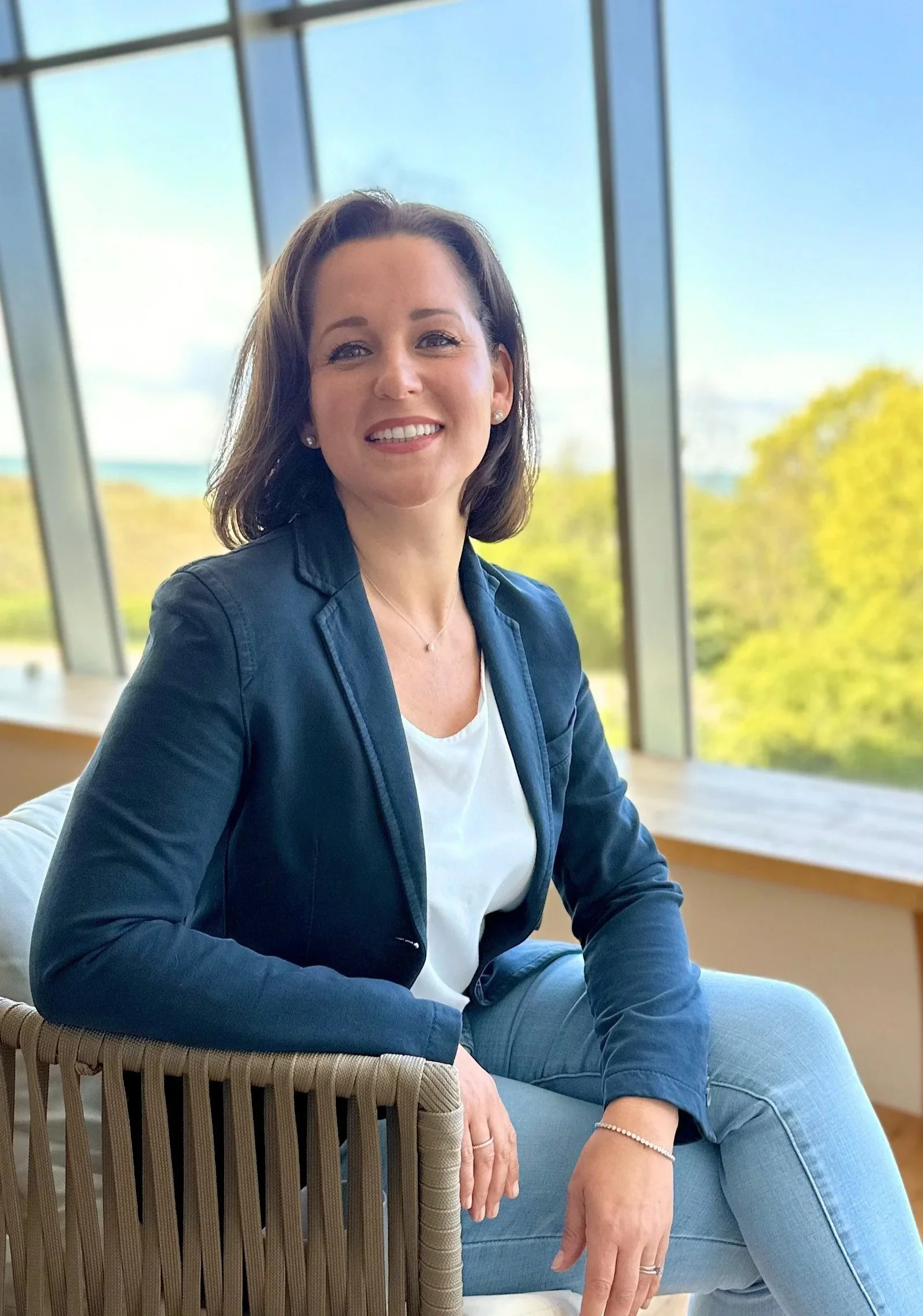 Eine lachende Frau sitzt in einem hellen Raum vor großen Fenstern mit Blick ins Grüne im Hintergrund, trägt eine blaue Jacke und Jeans, hat kurze braune Haare.