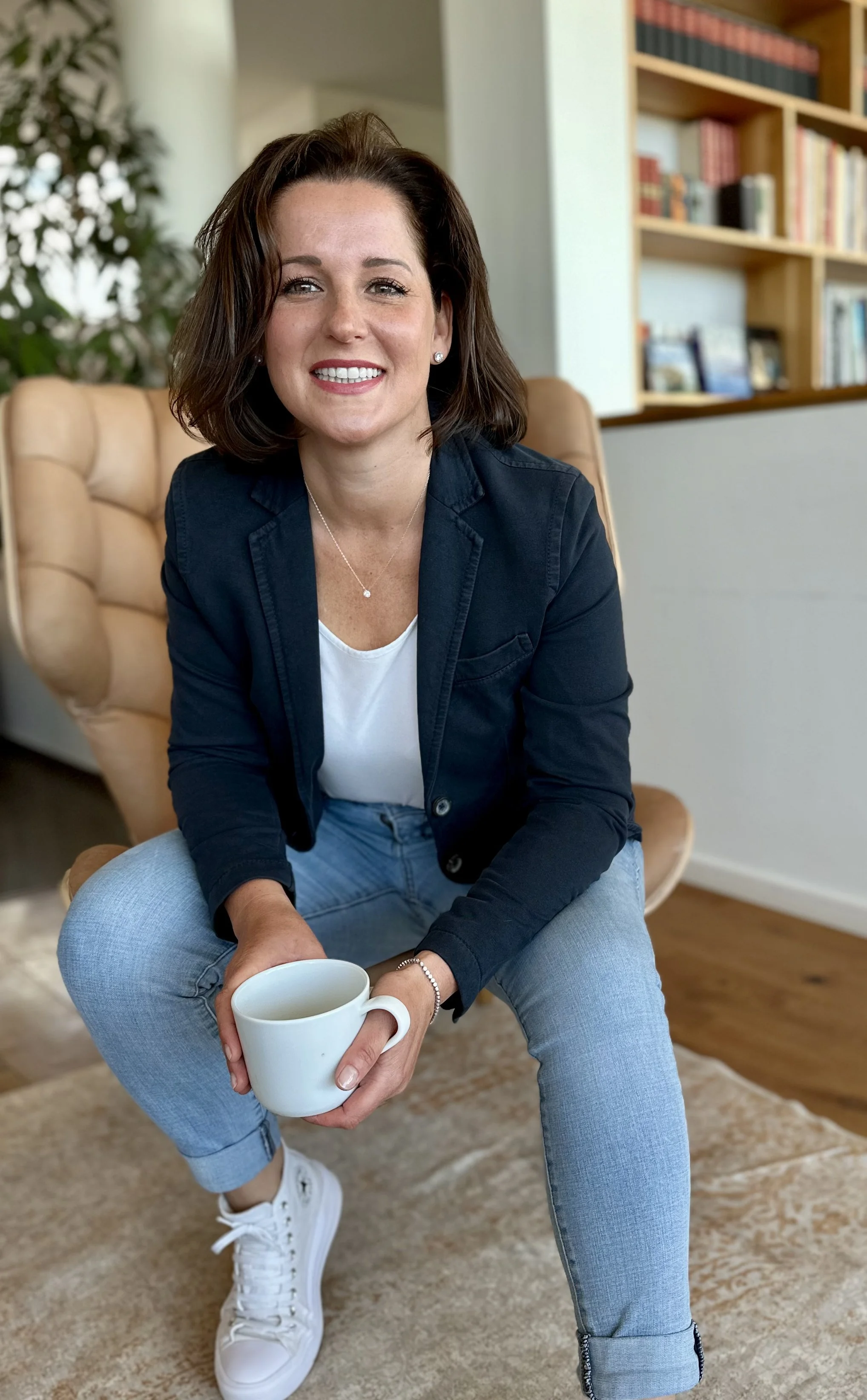 Eine lächelnde Frau sitzt bequem in einem Wohnraum, hält eine weiße Kaffeetasse in den Händen, trägt eine schwarze Jacke, ein weißes T-Shirt, hellblaue Jeans und weiße Turnschuhe, im Hintergrund ein Bücherregal.