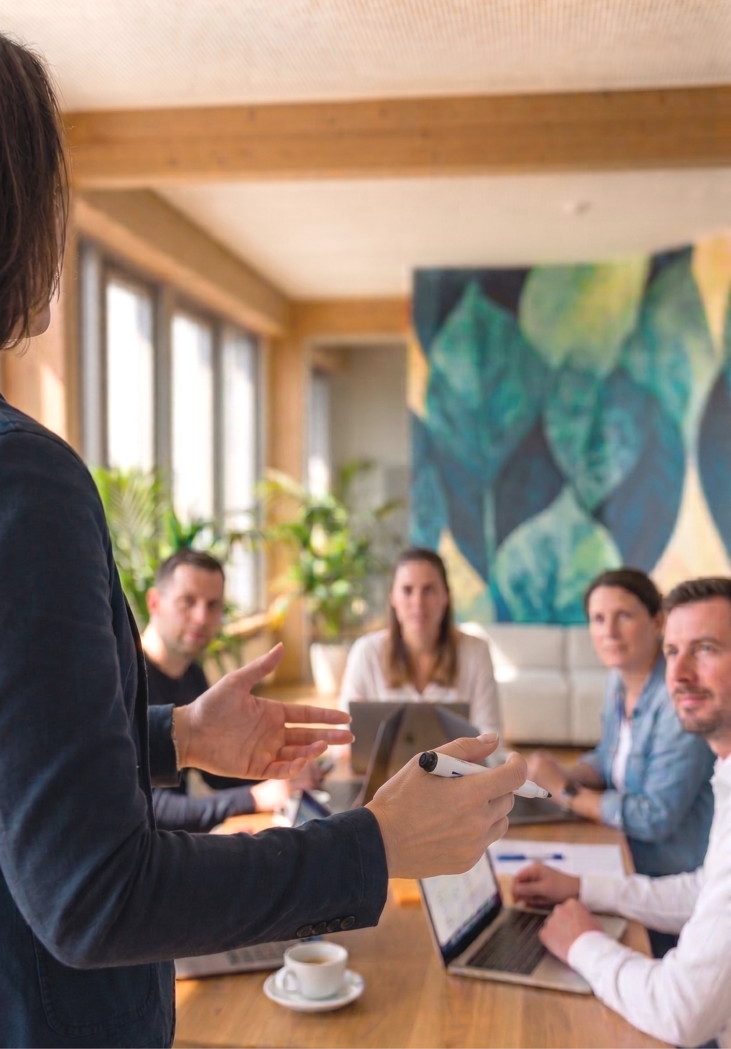 Menschen bei einer Besprechung, einer spricht und zeigt mit den Händen, andere mit Laptops und Tablets, im Raum gibt es Pflanzen und ein Gemälde an der Wand.