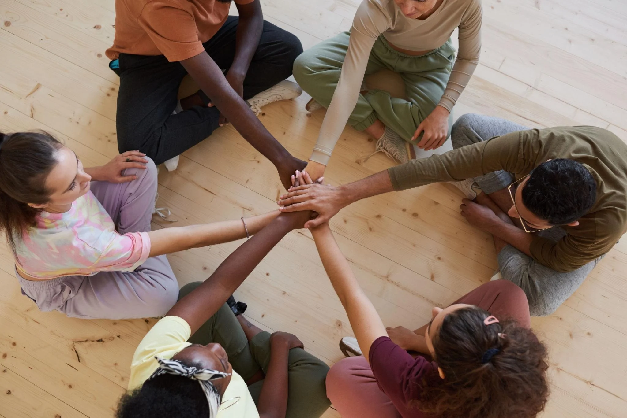A diverse group of people sitting on a wooden floor in a circle, with their hands stacked together in the center, symbolizing unity and teamwork.