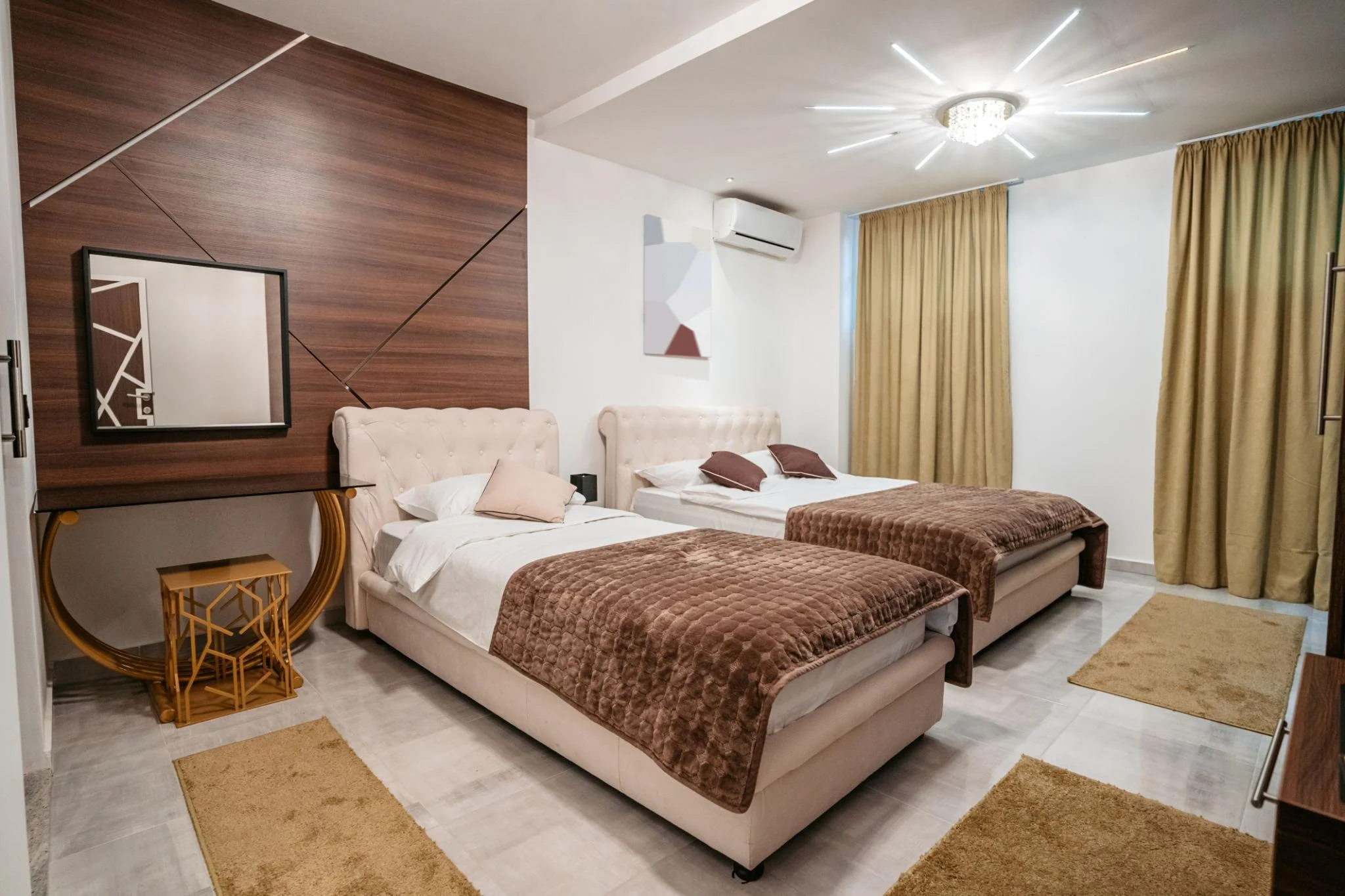 Hotel room with two beds, beige headboards, brown blankets, beige curtains, a wall-mounted mirror, a small side table, and modern ceiling light fixture.