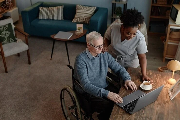 An elderly man in a wheelchair using a laptop, assisted by a caregiver in a living room with a teal sofa and wooden table.