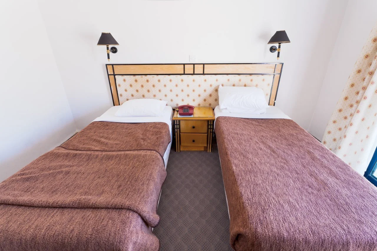 Two single beds with brown blankets and white pillows in a hotel room, separated by a nightstand with a red telephone, beige headboard with a floral pattern, and wall-mounted black lamps.