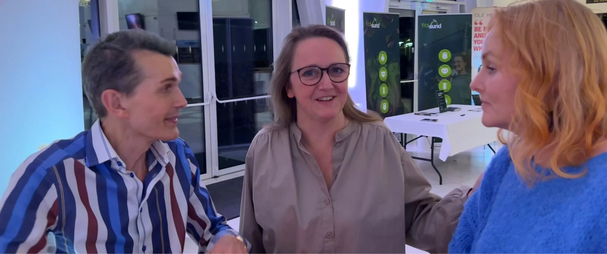 Tre personer taler og smiler i en række, mens de står i et indendørs område med informationsskærme og en bord med computer.