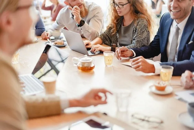 Mennesker i forretningsmøde, der sidder ved et langt bord med laptops, kopper, og drikkevarer.