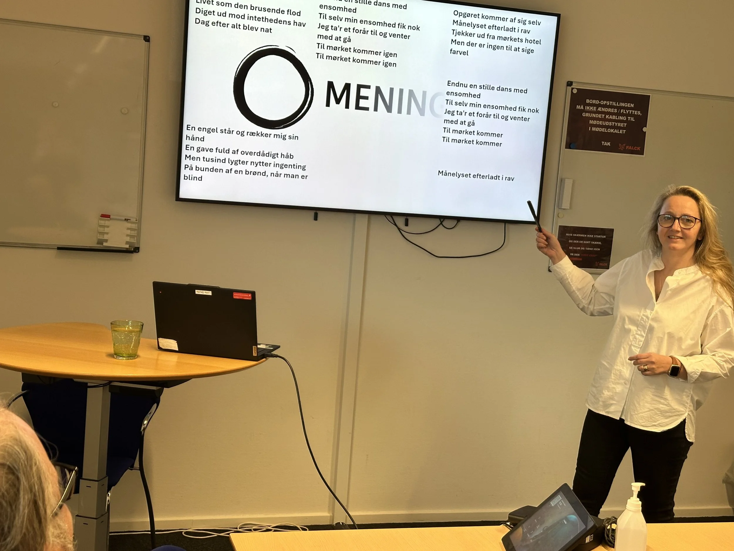 Kvinde står ved en tavle og peger på en stor skærm med tekst, mens hun fortæller. Der er et bord med en sort computer og et glas på, og en person i forgrunden ser mod præsentationen.