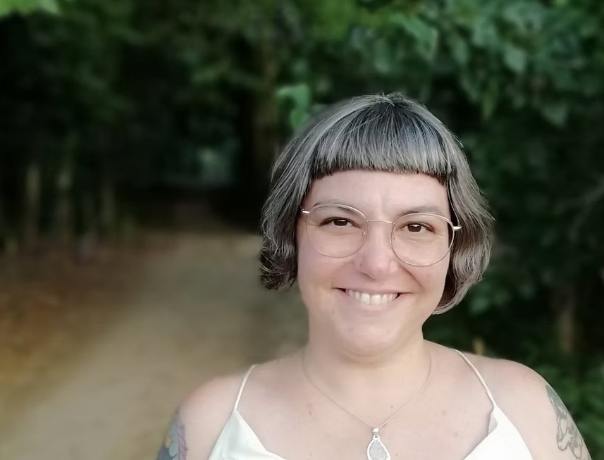 Mujer con gafas cortas de cabello, sonriendo en un entorno natural con árboles y camino de tierra.